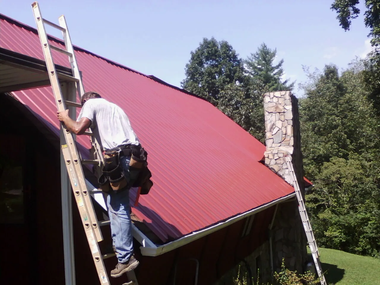 Roofer on a ladder beside a red metal roof and stone chimney — Soffit & Fascia Repair by Roofing Brothers in Bourneville